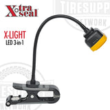 The image shows a black Xtra Seal X-Light LED 3-in-1 tire repair work light with a flexible neck and a clip-on handle, displayed against a white background. 14-923